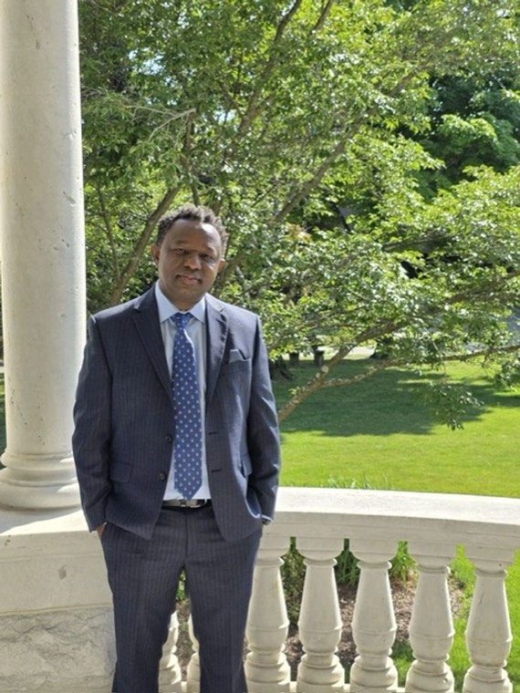 Charles Kangethe, candidate for Wawayanda Town Supervisor, standing on a porch in a dark suit and blue tie